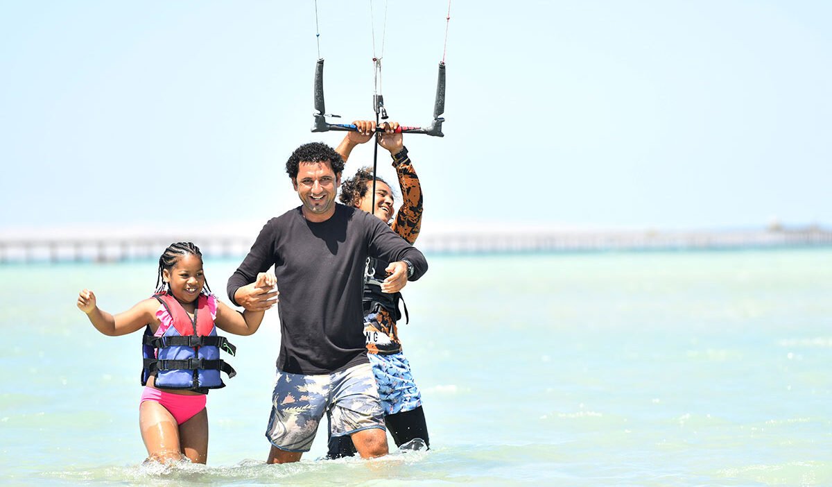 Beginners practicing kitesurf safety quick-release activation on a sandy beach.